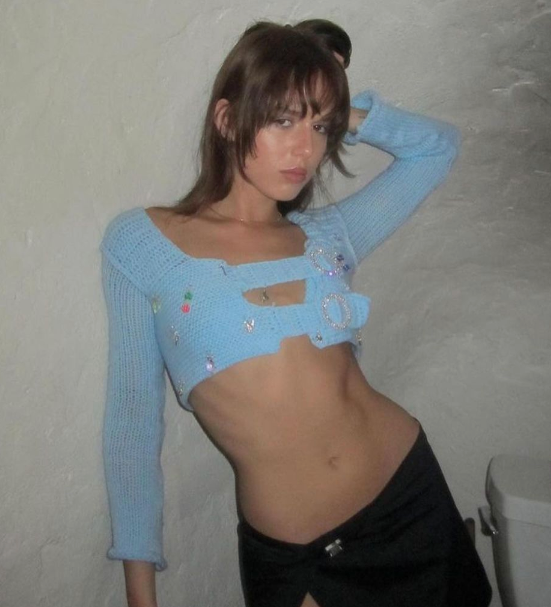 Model wearing the Bailey Prado Blue Long Sleeve Charm Top, a baby blue crochet crop top with rhinestone buckles, long sleeves, and colorful charm embellishments.