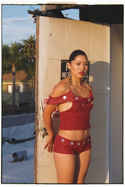 Woman in Handmade red knit seashell top and shorts set with shell embellishments by Bailey Prado standing against a wall with a palm tree in the background